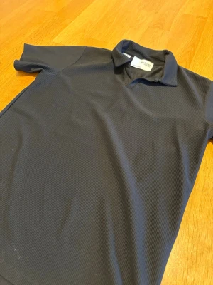 Mörkblå ribbad polotopp - Snygg mörkblå polotopp med korta ärmar och diskret ribbad struktur. Klassisk krage och enkel design som passar till mycket. Perfekt för dig som gillar stilrena plagg med en twist.