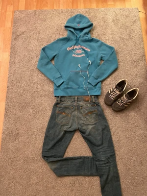 Blå Åre Peak Performance hoodie - Snygg blå Åre hoodie från Peak Performance med tryck i vitt och rött på bröstet där det står 'Peak Performance est. 1986 Are Adventurous'. Hoodien har huva med dragsko, magficka och ribbade muddar. Perfekt för en laidback, grisch look. Storlek = Large men själv skulle jag säga Medium. Säljer även Nudie Jeansen.
