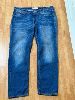 Blå raka jeans med fem fickor - Säljer ett par klassiska blå jeans med rak passform och fem fickor. Jeansen har snygga slitningar framtill och är tillverkade i denim. Perfekta för en avslappnad och trendig look.