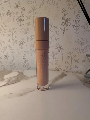 Flytande concealer med beige ton - Heltäckande Flytande concealer i en genomskinlig plasttub med beige lock. Produkten har en ljusbeige färg och kommer med applikator i locket. Förpackningen är smidig att ta med och har tryckt text i vitt. Passar perfekt för att täcka mörka ringar och ojämnheter.