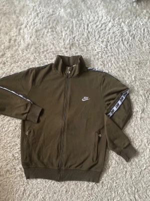 Olivgrön Nike ziptröja med logga - Olivgrön ziptröja från Nike med vit logga på bröstet och vita band med Nike-tryck längs ärmarna. Tröjan har hög krage, muddar vid ärmslut och nederkant samt fickor med dragkedja. Perfekt för en sportig och avslappnad stil.