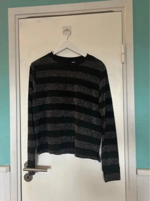 Svart tröja med glittriga ränder - Snygg svart långärmad tröja med breda, glittriga silverränder. Tröjan har rund halsringning och avslappnad passform. Perfekt för dig som gillar en enkel men ändå lite extra look.