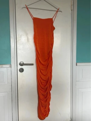 Orange långklänning med drapering - Snygg orange långklänning med smala axelband och draperad effekt längs hela kjolen. Klänningen har en figurnära passform och är perfekt för dig som vill sticka ut. Materialet känns lätt och följsamt.
