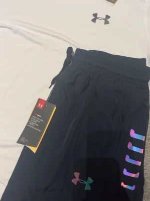 Under Armour träningsset svart/vit - Snyggt träningsset från Under Armour med en vit t-shirt och svarta byxor. Båda plaggen har färgglada, reflekterande loggor och detaljer. Byxorna har dragsko i midjan och elastiska muddar. Perfekt för gymmet eller löprundan.
