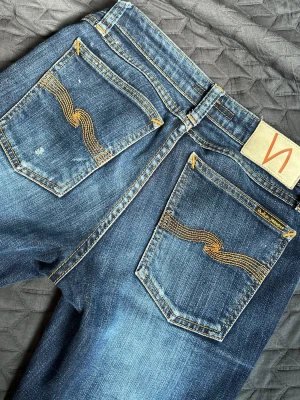 Blå nudie jeans - Snygga blå salvage nudie jeans med slitningar på knäna och låret. Jeansen har  klassiska bakfickor med dekorativa sömmar och är perfekta för en cool och avslappnad archive stil. 