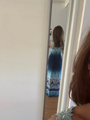 Blå långklänning med mönster - Säljer en blå långklänning med snyggt mönster och broderade detaljer vid axlarna. Klänningen har ett ombre-aktigt färgskifte från ljusblått till mörkblått och ett grafiskt mönster längst ner. Perfekt för sommarens alla tillfällen!