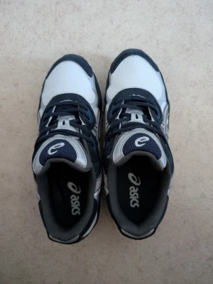 Asics - Snygga sneakers från Asics i vitt och svart med grå detaljer. Ovandelen är i mesh och syntetmaterial för bra andningsförmåga och komfort. Klassisk snörning och sportig design som passar till många olika stilar. Använd några gånger.