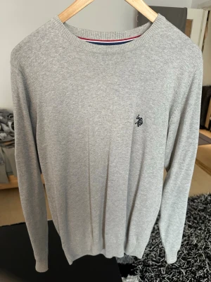 Ljusgrå stickad tröja U.S. Polo Assn. - Snygg ljusgrå stickad tröja från U.S. Polo Assn. med rund halsringning och diskret logga broderad på bröstet. Tröjan har långa ärmar och ribbade muddar vid ärmslut och nederkant. Perfekt till jeans eller chinos.