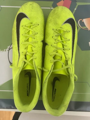 Nike Mercurial gula fotbollsskor - Säljer ett par Nike Mercurial fotbollsskor i neon-gult med svart swoosh på sidan. Skorna har snörning och låg profil, samt detaljer i svart och silver på hälen. Tillverkade i syntetmaterial för bra grepp och känsla på planen. Skorna är i pro en nivå under elites. De kostade mig 1000 kr och jag säljer de för halva pris eftersom de va för stora efter jag testat skorna och spelat med de bara en gång så de är i väldigt bra skick.