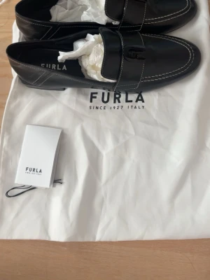 FURLA 1927 Loafers  - Stilrena svarta loafers från Furla i blankt skinn med dekorativ rem över foten och vita kontrastsömmar. Klassisk design med låg klack och rundad tå. Perfekta för dig som gillar en tidlös och elegant look.