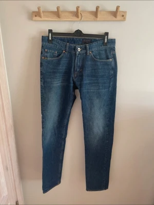 Blå raka jeans med slitningar - Säljer ett par snygga blå jeans från Tiger of Sweden. Klassisk femficksmodell med orangea sömmar och knappgylf. Jeansen har en cool tvättad look och passar perfekt till en avslappnad stil. Passformen är straight fit och storleken är true to size (29/32). Använda ett fåtal gånger, som nya!   Kontakta gärna för frågor!