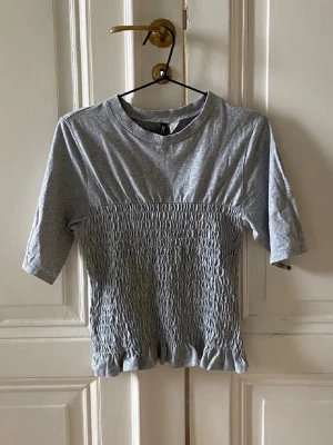 Grå topp med smock från HM - Grå topp från Divided med smockad midja och volangkant nedtill. T-shirten har rund halsringning och korta ärmar. Tillverkad i mjuk bomull som känns skön mot huden. Enkel men ändå unik detalj med smock och volang.
