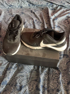 Nike skor - Snygga svarta Nike sneakers med vit sula och vit swoosh på sidan. Skorna har lågt skaft, svarta skosnören och ovandel i textil och syntet. Perfekta för en sportig och avslappnad stil.
