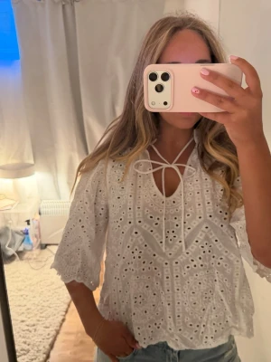 Vit broderad blus med knytning - Säljer en vit blus med broderade detaljer och hålmönster. Blusen har trekvartsärmar, v-ringning med knytband och vågig kant nedtill. Perfekt till jeans eller kjol för en fräsch look. Materialet känns lätt och luftigt.