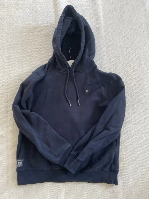 Mörkblå Morris hoodie - Snygg mörkblå hoodie med huva. Liten broderad Morris logga på bröstet och en patch nertill vid midjan. Tillverkad i mjukt bomullsliknande material och har ribbade muddar vid ärmar och nederkant. Perfekt för en avslappnad och stilren look.