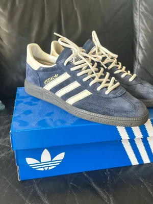 Adidas Spezial blåa sneakers - Snygga Adidas Spezial sneakers i mörkblå mocka med vita detaljer och klassiska tre ränder på sidan. Skorna har vita snören och en mörk sula i gummi. Perfekta för dig som gillar retrostil och streetwear.
