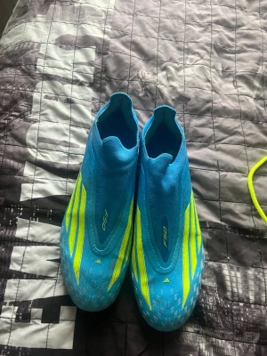 Blåa Adidas F50 fotbollsskor - Säljer ett par blåa Adidas F50 fotbollsskor med gula detaljer och klassiska tre ränder på sidan. Skorna har en strömlinjeformad design utan snörning och är tillverkade i ett lätt syntetmaterial för maximal fart på planen. 