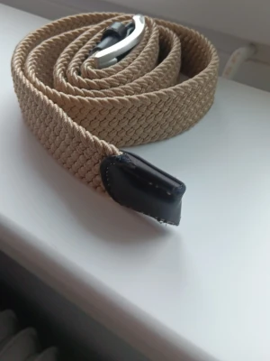 Beige flätat bälte med skinn-detaljer - Snyggt beige flätat bälte med silverfärgat spänne och svarta skinn-detaljer vid ändarna. Perfekt accessoar för att lyfta din outfit och funkar till både jeans och chinos. Bältet har en stilren och modern look.