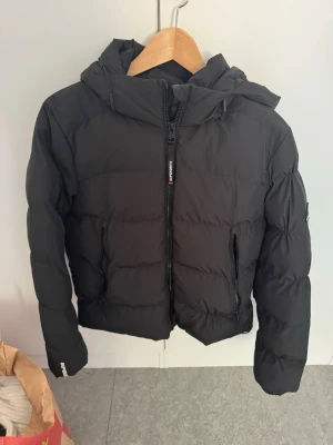 Svart pufferjacka med huva - Säljer en svart pufferjacka med huva och dragkedja framtill. Jackan har två sidofickor med dragkedja och en sportig look. Perfekt för kalla dagar och riktigt skön att bära. Passar dig som vill ha en stilren och enkel vinterjacka.