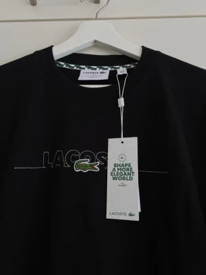 Lacoste T-shirt - Oanvänd Lacoste T-shirt i 100 % bomull. Tyvärr tog jag bort prislappen eftersom jag inte planerade att sälja t-shirten tillsammans med den. Tog dock en bild på den precis innan. Jag är 180 cm lång och väger 65 kg. T-shirten passar mig perfekt men skulle även kunna passa längre eller större personer. Fick den i present men anser inte att det är min stil.