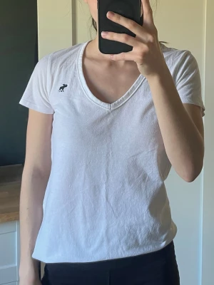 Vit v-ringad t-shirt Abercrombie - Klassisk vit t-shirt från Abercrombie & Fitch med v-ringning och korta ärmar. T-shirten har en liten mörkblå logga broderad på bröstet och är tillverkad i mjuk bomull. Perfekt basplagg med enkel och stilren design.