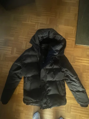 Svart canada goose vinter jacka - Säljer en svart pufferjacka med stor, mysig huva och breda kanaler. Jackan har dragkedja och knappar framtill samt två sidofickor. Perfekt för kalla vinterdagar och riktigt skön att bära. Materialet är glansigt och ger ett modernt intryck.