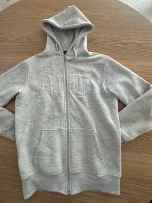 Tuttolente  - Storlek XS. Snygg ljusgrå hoodie med dragkedja och huva. På framsidan finns broderade bokstäver 'M' och 'T' i gotisk stil och på ryggen ett diskret tryck av en örn. Hoodien har känguruficka och är gjord i mjukt sweatshirtmaterial.