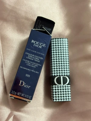 Rött Rouge Dior läppstift 999 - Rouge Dior läppstift i nyansen 999 med elegant houndstooth-mönstrad hylsa och silverdetaljer. Läppstiftet har en intensiv röd färg och klassisk rundad form. Lyxig förpackning och ikonisk Dior-logga på hylsan.
