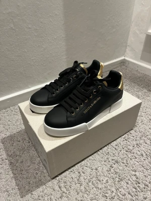 Svarta Dolce & Gabbana sneakers - Snygga svarta sneakers från Dolce & Gabbana med gulddetaljer på häl och snörhål. Skorna har en vit tjock sula och är tillverkade i slätt läder. Klassisk låg modell med svart snörning och diskret logga i guld på sidan. Nypris:7000kr mitt pris: 3000kr