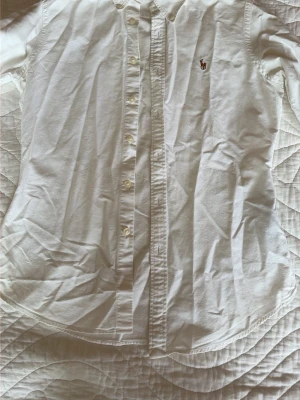 Vit skjorta med broderad logga - Klassisk vit skjorta i bomull med knappar framtill och en liten broderad logga på bröstet. Skjortan har lång ärm och tidlös design som passar till många olika stilar. 