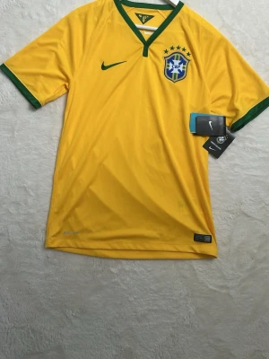 Gul Brasilien fotbollströja Nike - Snygg gul fotbollströja från Nike med gröna detaljer och Brasilien-emblem på bröstet. Tröjan har V-ringning med grön kant och korta ärmar med gröna muddar. Perfekt för dig som älskar fotboll och vill stötta Brasilien.