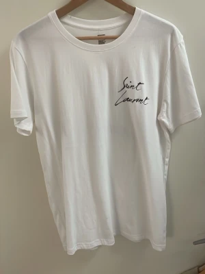 L passar M - Snygg vit t-shirt med svart Saint Laurent-tryck både fram och bak. Klassisk rund hals och korta ärmar. Perfekt för en stilren och trendig look. Tillverkad i mjuk bomull som känns skön mot huden.