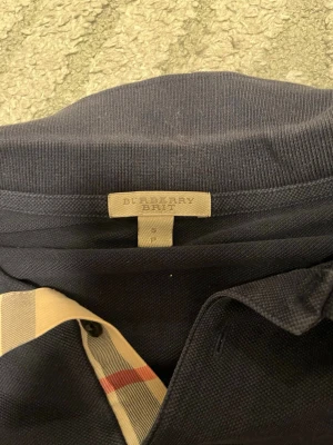 Mörkblå pikétröja från Burberry Brit - Säljer en mörkblå pikétröja från Burberry Brit med klassisk krage och korta ärmar. Tröjan har Burberrys ikoniska rutiga detalj vid knappslån och en liten broderad logga på bröstet. Perfekt för en stilren och avslappnad look.
