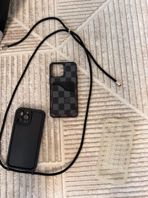 Tre mobilskal iPhone 12 Pro Max  - Säljer tre olika mobilskal: ett svart skal med snodd, ett rutigt skal i svart och grått med kortfack och röd söm, samt ett transparent skal med bubbelmönster. Passar dig som vill variera stilen på din mobil och gillar både klassiskt och lekfullt.