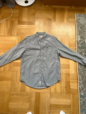 Grå skjorta från Timberland - Säljer en stilren grå skjorta från Timberland med button-down krage och långa ärmar. Skjortan har en bröstficka med Timberland-logga broderad ovanför fickan. Perfekt till jeans eller chinos för en clean look.