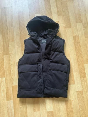 Svart dunväst med huva från ZARA - Svart dunväst från ZARA med stor huva och två rejäla fickor framtill. Västen är vadderad och har en stilren, rak passform. Perfekt för lager-på-lager och enkel att matcha med olika outfits.