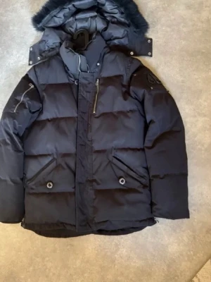 Svart dunjacka Moose Knuckles - Snygg svart dunjacka från Moose Knuckles med avtagbar huva och pälskant. Jackan har flera fickor med dragkedja och knappar, samt en logga på ärmen. Perfekt för kalla vinterdagar tack vare tjockt dunfoder och vindtätt material.