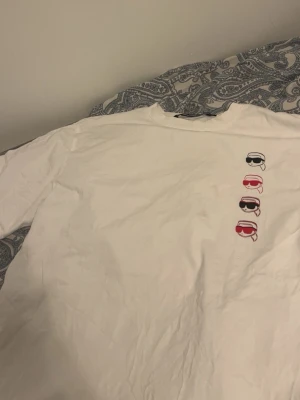 Vit Karl Lagerfeld t-shirt med tryck - Säljer en vit t-shirt från Karl Lagerfeld med coolt tryck av fyra små figurer med solglasögon i svart och rött på bröstet. Klassisk passform och rund hals. Perfekt för dig som gillar stilrena plagg med en twist.