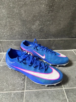 Blå Nike Mercurial fotbollsskor - Säljer ett par blå Nike Mercurial fotbollsskor med vita och rosa detaljer. Skorna har snörning, låg profil och är tillverkade i ett lätt syntetmaterial för bästa känsla på planen. Perfekta för dig som vill ha speed och kontroll på gräs.