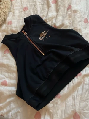 Svart croppad Nike Air linnetopp - Snygg svart croppad linnetopp från Nike Air med halv dragkedja framtill och logga i silver. Perfekt för en sportig look och passar till både jeans och träningsbyxor.