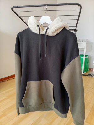 Svart och olivgrön hoodie från Denim Day - Säljer en oversized hoodie från Denim Day i svart och olivgrönt med stor magficka och mysig teddyfodrad huva. Hoodien har dragsko i huvan och är tillverkad i mjukt material, perfekt för chill dagar. Färgerna är stilrena och passar till det mesta.