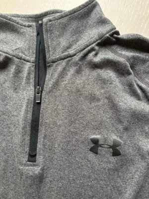 Grå halvzip tröja från Under Armour - Mjuk och skön grå långärmad tröja från Under Armour med halvzip framtill. Tröjan har hög krage och är tillverkad i ett stretchigt material som passar perfekt för träning eller chill. Diskret logga på bröstet och enkel, stilren design.
