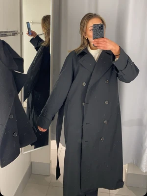 Svart oversized trenchcoat - Stilren svart trenchcoat med dubbelknäppning och bred krage. Modellen är oversized med långa ärmar och ett matchande knytbälte i midjan. Klassisk design som passar perfekt till lager-på-lager och ger en cool, avslappnad look.