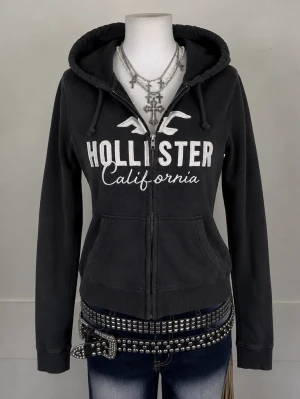 Svart Hollister hoodie med dragkedja - Snygg svart hoodie från Hollister med vit logga och texten 'California' på bröstet. Hoodien har dragkedja framtill, två fickor och justerbar huva med snören. Tillverkad i mjukt bomullsmaterial som är skönt att bära.
