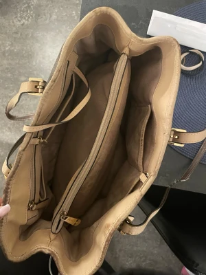 Brun Michael Kors handväska - Stilren brun handväska från Michael Kors med beigea detaljer och guldfärgade spännen. Väskan har flera fack, dragkedja i mitten och ett klassiskt MK-hänge. Tillverkad i tåligt konstläder med logomönster över hela ytan. Perfekt för dig som vill ha plats för allt.