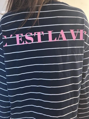 Randig långärmad topp med text - Mörkblå långärmad topp med vita horisontella ränder och rosa texten 'C'EST LA VIE' på ryggen. Oversized passform och mjukt material som känns skönt mot huden. Perfekt för en avslappnad och trendig look.