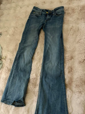 Blå bootcut jeans med låg midja - Säljer ett par klassiska blå bootcut jeans i denim med låg midja och fem fickor. Jeansen har en lätt tvättad look och vida ben från knät och ner. Perfekta för dig som gillar retrostil och vill ha ett par sköna jeans till vardags.