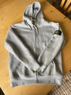 Grå zip-hoodie med patch på ärmen - Snygg grå hoodie med dragkedja och huva från Stone Island. Hoodien har kängurufickor och ribbade muddar. Perfekt för en avslappnad streetwear-look. Storleken är M men passar som S också bara lite mer avslappnad passaform så ifall man gillar lite mer baggy relaxed fit så är den perfekt. Hör gärna av dig vid funderingar.