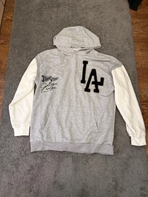 Grå och vit hoodie från LA - Snygg hoodie från LA i grått med vita ärmar. Stor LA-logga i svart på bröstet och tryck med texten 'Champ Team College League'. Mjuk huva och känguruficka framtill. Perfekt för en chill och sportig stil.