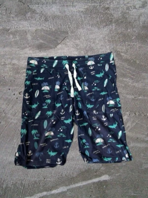 Mörkblå shorts med strandmönster - Säljer ett par mörkblå shorts med snörning i midjan och coolt strandinspirerat mönster med palmer, hajar, surfbrädor och ankare. Perfekta för sommaren och strandhäng. Lätta och bekväma att bära.